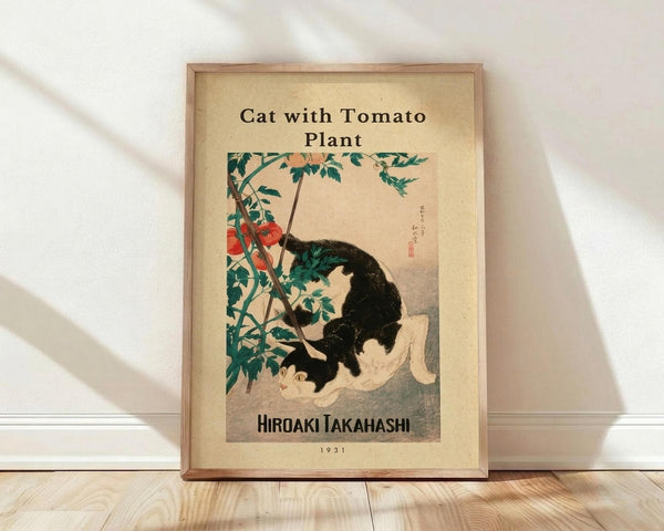 May include: Framed vintage Japanese woodblock print showing a black and white cat sitting beside a tomato plant with red tomatoes and green leaves. The artwork has aged beige paper background with Japanese text and artist signature. Wooden frame leans against white wall on light wooden floor with soft natural lighting creating diagonal shadows.
