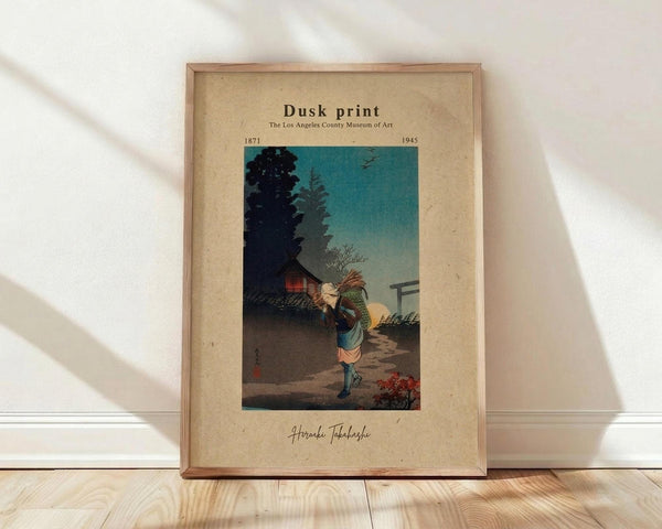 May include: A framed vintage Japanese woodblock print showing a figure carrying bundles of sticks along a twilight path, with traditional buildings and pine trees silhouetted against a turquoise evening sky. The artwork is displayed in a light wood frame with cream matting, leaning against a white wall on wooden flooring with natural window light casting shadows.