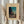May include: Hands holding a framed vintage Japanese woodblock print showing a figure carrying bundles at dusk, with teal sky and traditional buildings. Cream matted frame against white curtained window. Dried wheat stems held in left hand. Person wearing beige ruffle-sleeve top.