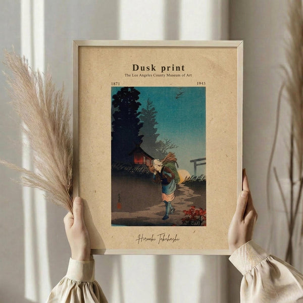 May include: Hands holding a framed vintage Japanese woodblock print showing a figure carrying bundles at dusk, with teal sky and traditional buildings. Cream matted frame against white curtained window. Dried wheat stems held in left hand. Person wearing beige ruffle-sleeve top.