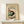 May include: Framed Japanese woodblock-style art print showing a black and white cat beneath a tomato plant with red fruit and green leaves, displayed on a light wood shelf beside a small wrapped gift with evergreen sprig. The print has cream background with brown text and is set in a natural wood frame against a white wall.