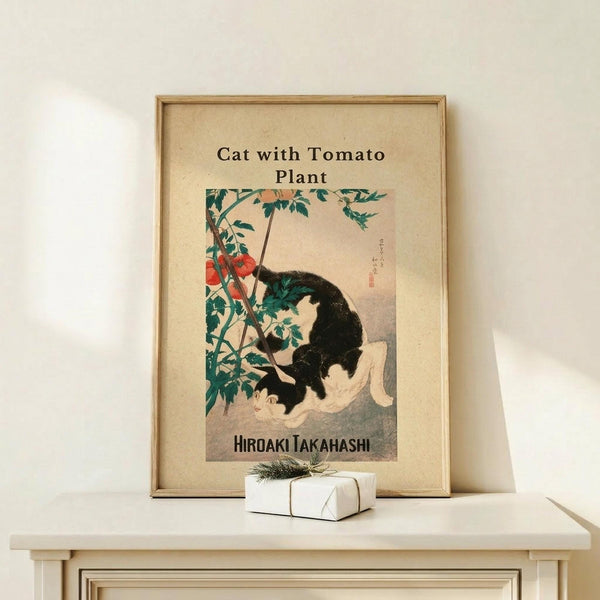 May include: Framed Japanese woodblock-style art print showing a black and white cat beneath a tomato plant with red fruit and green leaves, displayed on a light wood shelf beside a small wrapped gift with evergreen sprig. The print has cream background with brown text and is set in a natural wood frame against a white wall.