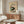 May include: Japanese woodblock-style poster print featuring black and white cat with tomato plant, mounted on cream-colored wall above walnut wood credenza. Modern living room setting with white curved chair, glass oval coffee table, and minimalist decor. Print shows vintage aesthetic with green foliage and red tomatoes against beige background.