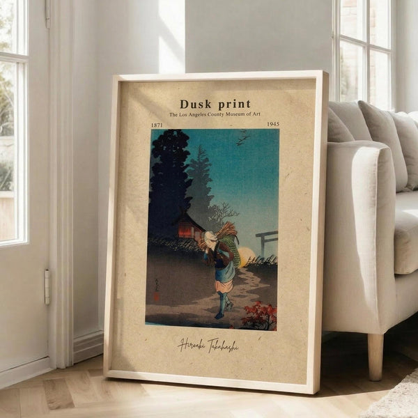May include: Framed vintage-style museum poster featuring Japanese woodblock print of person carrying bundles of sticks at dusk, with teal sky and silhouetted trees. Cream matting with museum attribution text and artist signature. Wooden frame leaning against white wall next to beige upholstered chair in bright, minimalist living room with large windows and natural lighting.