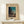 May include: Framed vintage Japanese woodblock print showing a figure carrying bundles of wood or straw along a twilight path, with traditional buildings and trees silhouetted against a turquoise-green evening sky. The print is displayed in a light wooden frame on a cream-colored wooden surface alongside a small white gift box tied with twine and a sprig of greenery. Warm, minimalist styling with neutral tones throughout the scene.