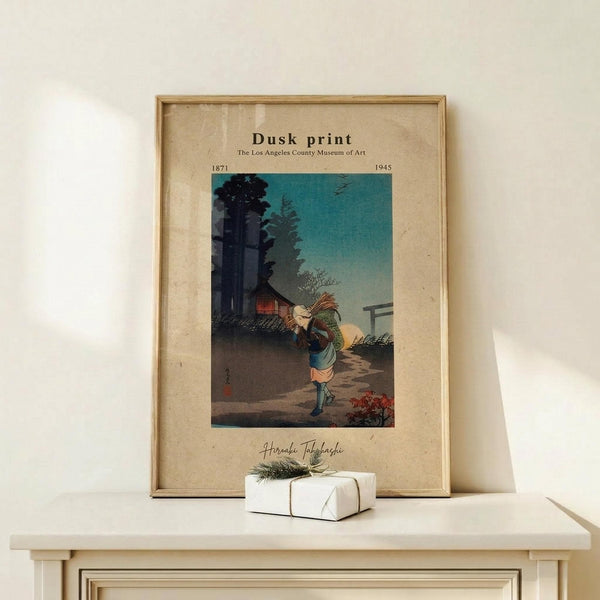 May include: Framed vintage Japanese woodblock print showing a figure carrying bundles of wood or straw along a twilight path, with traditional buildings and trees silhouetted against a turquoise-green evening sky. The print is displayed in a light wooden frame on a cream-colored wooden surface alongside a small white gift box tied with twine and a sprig of greenery. Warm, minimalist styling with neutral tones throughout the scene.
