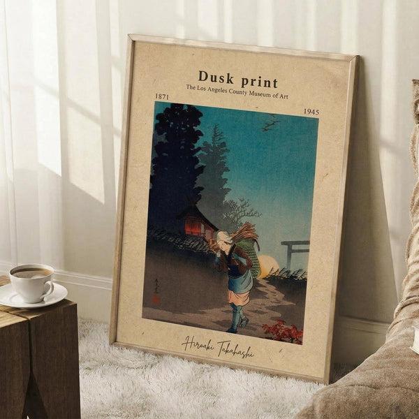 May include: Framed vintage Japanese woodblock print showing a figure carrying bundles walking at dusk, with traditional buildings and trees silhouetted against a turquoise evening sky. The beige matted frame leans against white wainscoting near a wooden side table with coffee cup and textured throw blanket on white carpet.