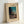 May include: Framed vintage Japanese woodblock print showing a figure in traditional clothing carrying bundles of wood at dusk, with turquoise evening sky, dark silhouetted trees, and a small house with glowing window. Print has cream colored mat with museum text and artist signature, displayed in light wood frame leaning against white wall.