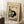 May include: Framed vintage-style poster leaning against white wall corner on textured cream rug. Features black and white cat sitting beside tomato plant with red tomatoes and green foliage on cream background. Japanese characters visible on artwork. Coffee cup on wooden block nearby, beige blanket partially visible.