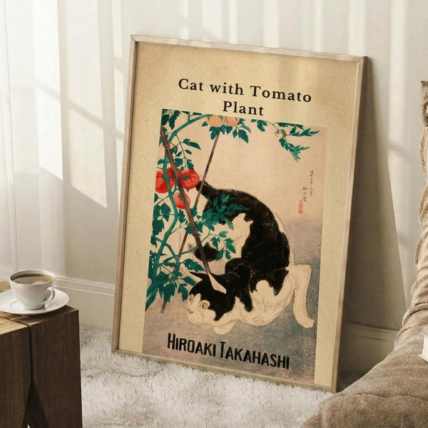 May include: Framed vintage-style poster leaning against white wall corner on textured cream rug. Features black and white cat sitting beside tomato plant with red tomatoes and green foliage on cream background. Japanese characters visible on artwork. Coffee cup on wooden block nearby, beige blanket partially visible.