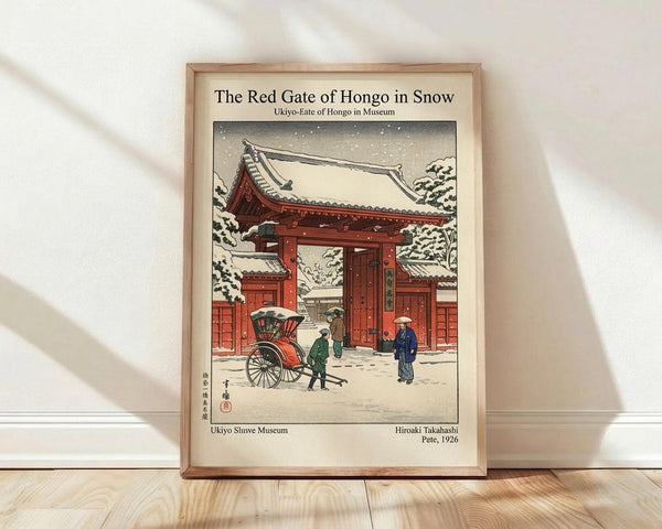 May include: Framed vintage Japanese woodblock print showing a red temple gate in falling snow, with people and a rickshaw in the courtyard. The artwork features traditional architecture with snow-covered roofs, figures in winter clothing, and muted colors against a snowy landscape. The print is displayed in a light wooden frame, leaning against a white wall on wooden flooring with soft natural lighting.