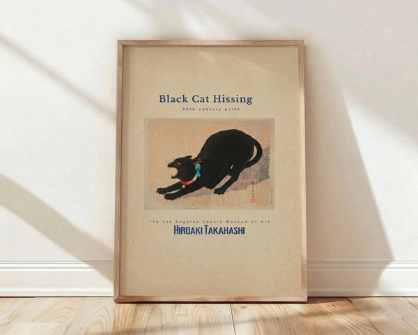 May include: Framed vintage Japanese woodblock print poster featuring a black cat in an arched, hissing pose with blue and red collar details against a peachy background. The cream-colored poster displays museum text and artist name below the artwork. Natural wood frame leans against white wall on light wooden floor with soft window light casting shadows.