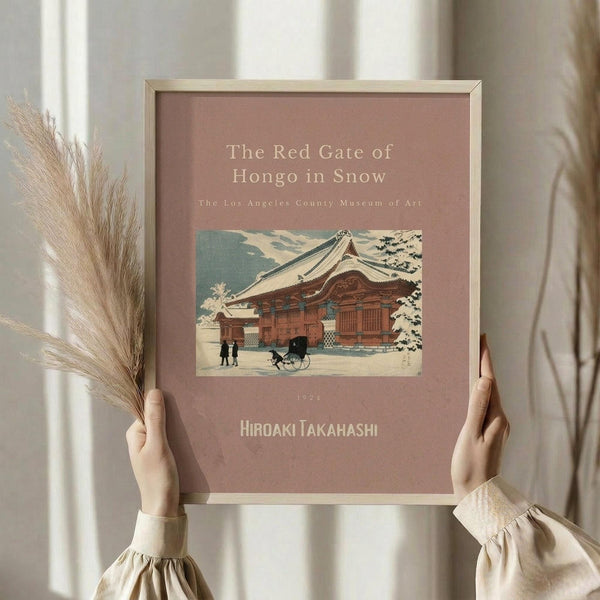 May include: Hands holding a framed vintage Japanese woodblock print showing a traditional red temple gate in snow. The artwork features muted colors with a pink-beige mat and thin wooden frame. Dried pampas grass stems are visible to the left, with soft natural lighting and neutral curtains in the background creating a minimalist, cozy interior setting.