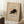 May include: Framed vintage Japanese woodblock print of a black cat in an arched, hissing pose with blue collar, displayed on cream matting in light wood frame. Poster leans against white wall corner on fluffy white rug beside wooden side table with white coffee cup and saucer. Soft natural lighting creates geometric shadows on wall. Cozy bedroom setting with neutral beige bedding visible at edge.