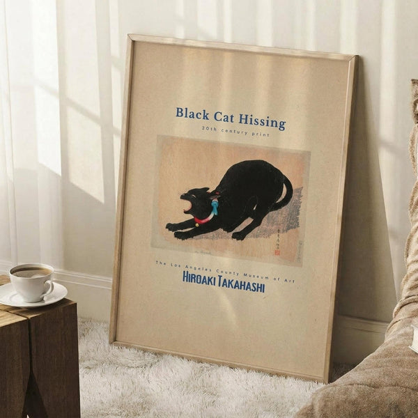 May include: Framed vintage Japanese woodblock print of a black cat in an arched, hissing pose with blue collar, displayed on cream matting in light wood frame. Poster leans against white wall corner on fluffy white rug beside wooden side table with white coffee cup and saucer. Soft natural lighting creates geometric shadows on wall. Cozy bedroom setting with neutral beige bedding visible at edge.