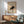 May include: A framed museum poster featuring a black cat in a stretching or hissing pose against a tan background, displayed above a wooden sideboard with sliding doors. The print has museum text and artist attribution. A mint green mid-century modern armchair with wooden frame sits beside the credenza, along with a white ceramic table lamp. Natural sunlight creates geometric shadows across the white wall and wooden furniture.