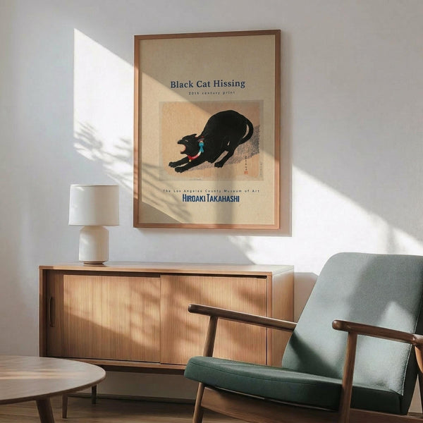 May include: A framed museum poster featuring a black cat in a stretching or hissing pose against a tan background, displayed above a wooden sideboard with sliding doors. The print has museum text and artist attribution. A mint green mid-century modern armchair with wooden frame sits beside the credenza, along with a white ceramic table lamp. Natural sunlight creates geometric shadows across the white wall and wooden furniture.