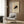 May include: Framed vintage Japanese art print featuring a black cat in an arched, hissing pose on cream background, displayed on wood-paneled wall above beige upholstered armchair with geometric patterned pillow, alongside brass floor lamp and dark marble planter in modern neutral-toned room setting.