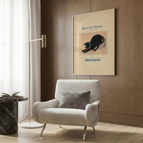 May include: Framed vintage Japanese art print featuring a black cat in an arched, hissing pose on cream background, displayed on wood-paneled wall above beige upholstered armchair with geometric patterned pillow, alongside brass floor lamp and dark marble planter in modern neutral-toned room setting.