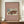 May include: Framed vintage Japanese woodblock print poster displayed on wooden sideboard beside white ceramic vase and books. The artwork shows a traditional red temple building covered in snow with small figures and cart in courtyard. Dusty rose background with cream text identifying the Los Angeles County Museum of Art and artist Hiroaki Takahashi, dated 1926. Thin dark wood frame against cream wainscoted wall.
