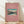 May include: Framed vintage Japanese woodblock print poster featuring Mt. Fuji with snow-covered peak rising above a serene lake scene with trees and small boats. The artwork has a pink coral background with turquoise and earth tones in the landscape. Natural wood frame leaning against white wall on light wooden floor with soft shadows.