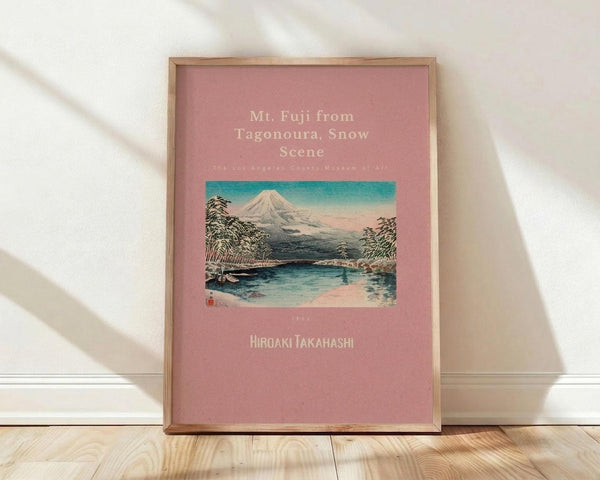 May include: Framed vintage Japanese woodblock print poster featuring Mt. Fuji with snow-covered peak rising above a serene lake scene with trees and small boats. The artwork has a pink coral background with turquoise and earth tones in the landscape. Natural wood frame leaning against white wall on light wooden floor with soft shadows.