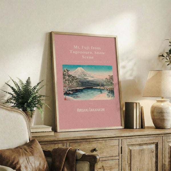 May include: Framed vintage Japanese woodblock print poster featuring Mt. Fuji with snow-capped peak, lake, and trees on pink background, displayed on rustic wooden console table alongside fern plant, books, ceramic vase, and cream upholstered furniture in neutral-toned living room setting.