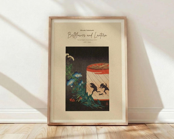 May include: Framed vintage Japanese woodblock print showing blue bellflowers and foliage beside a red paper lantern with two black silhouetted figures beneath it, displayed in light wood frame against white wall on wooden floor with natural window lighting.