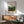 May include: Framed Henri Rousseau tropical forest poster print hanging above a wooden sideboard in a bright living room. The artwork shows dense green jungle foliage with red flowering plants and monkeys. Museum-style poster has cream background with brown wooden frame. Mid-century modern furniture includes sage green upholstered chair with wood arms and white ceramic table lamp. Natural sunlight casts shadows across white wall.