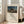 May include: Framed poster print leaning against white wall next to beige sofa in bright room with hardwood floors. Print shows Henri Rousseau's desert scene with sleeping figure and lion under moonlit sky, displayed in light wooden frame with cream matting and modern typography below the artwork.
