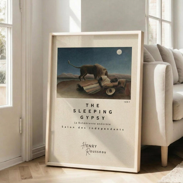 May include: Framed poster print leaning against white wall next to beige sofa in bright room with hardwood floors. Print shows Henri Rousseau's desert scene with sleeping figure and lion under moonlit sky, displayed in light wooden frame with cream matting and modern typography below the artwork.