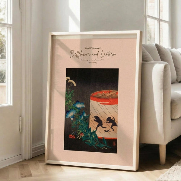 May include: Framed art print with pale pink matting in light wood frame, leaning against white wall next to beige sofa. The artwork shows Japanese-style illustration with blue morning glory flowers, green foliage, and black silhouettes of two figures near a red paper lantern against dark background. Natural sunlight streams through windows onto hardwood floors in bright, minimalist living room setting.