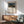 May include: Framed art print featuring a sleeping figure with a lion in a moonlit desert landscape, displayed above a wooden sideboard with white ceramic lamp in modern living room with mint green mid-century armchair and round coffee table, natural sunlight casting shadows across white wall