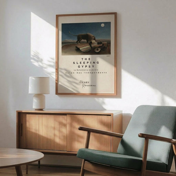 May include: Framed art print featuring a sleeping figure with a lion in a moonlit desert landscape, displayed above a wooden sideboard with white ceramic lamp in modern living room with mint green mid-century armchair and round coffee table, natural sunlight casting shadows across white wall