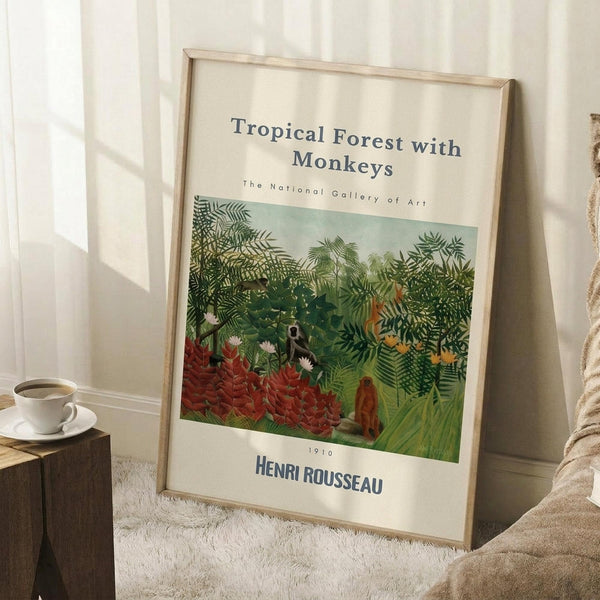 May include: Framed art print leaning against white wall showing lush tropical jungle scene with dense green foliage, red plants, and monkeys among the trees. Light wood frame, museum-style layout with cream matting and black text. Small wooden side table with white coffee cup nearby, beige throw blanket visible, soft natural lighting creating shadows on textured white rug.