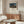 May include: A framed art print of Henri Rousseau's "The Sleeping Gypsy" hanging above a mid-century modern wooden credenza with sliding doors. The poster shows a lion approaching a sleeping figure under a full moon in a desert landscape. The room features a white curved chair, oval glass coffee table, brass floor lamp, and white table lamp, all styled in warm neutral tones with natural lighting creating soft shadows.