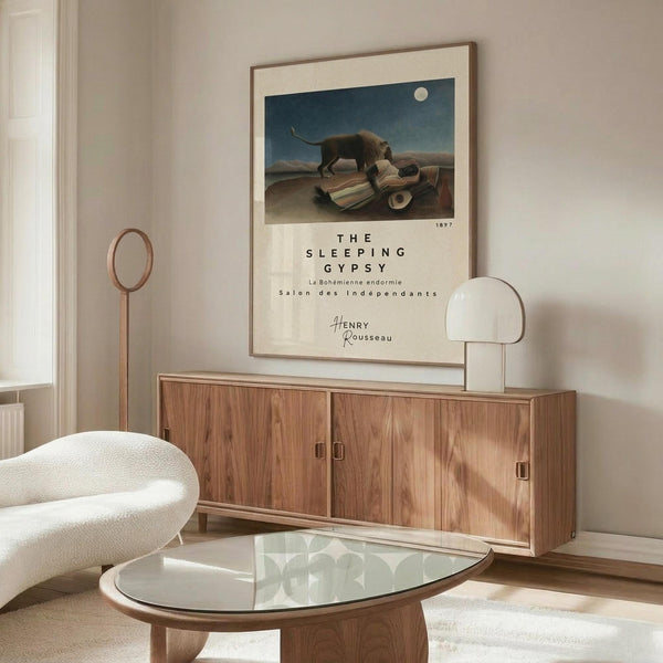 May include: A framed art print of Henri Rousseau's "The Sleeping Gypsy" hanging above a mid-century modern wooden credenza with sliding doors. The poster shows a lion approaching a sleeping figure under a full moon in a desert landscape. The room features a white curved chair, oval glass coffee table, brass floor lamp, and white table lamp, all styled in warm neutral tones with natural lighting creating soft shadows.