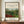 May include: Framed art print displaying a lush tropical jungle scene with dense green foliage, red flowering plants, and monkeys among the trees. The poster features cream-colored text areas above and below the botanical illustration, mounted in a thin brown frame. Displayed on a wooden sideboard alongside a small white ceramic vase and stacked books against white paneled walls.