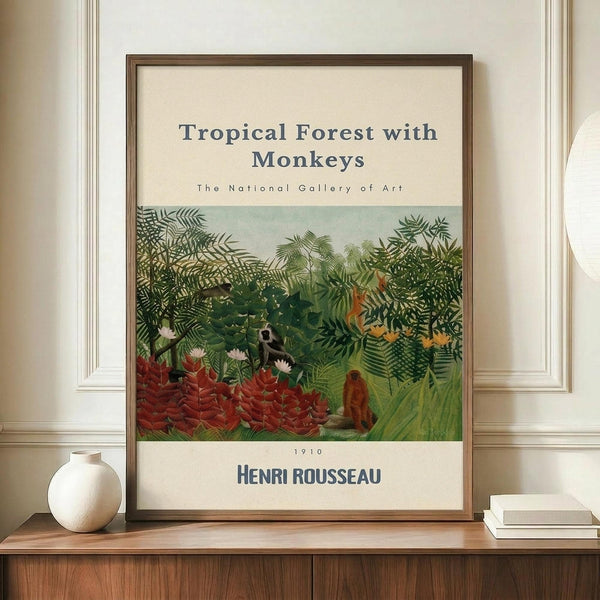 May include: Framed art print displaying a lush tropical jungle scene with dense green foliage, red flowering plants, and monkeys among the trees. The poster features cream-colored text areas above and below the botanical illustration, mounted in a thin brown frame. Displayed on a wooden sideboard alongside a small white ceramic vase and stacked books against white paneled walls.