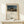May include: Framed poster print of Henri Rousseau's surreal desert painting featuring a sleeping figure in colorful robes beside a lion under a full moon. The reproduction shows muted earth tones and a dreamlike landscape. Displayed in a light wood frame on a white shelf next to a small wrapped gift with twine and greenery sprig against a cream wall.