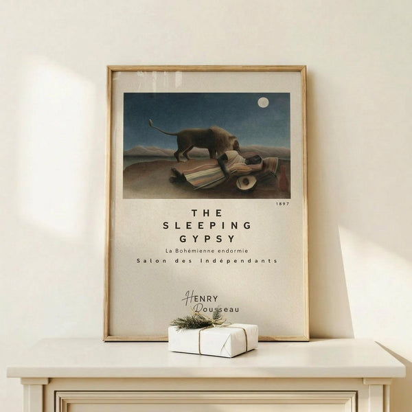 May include: Framed poster print of Henri Rousseau's surreal desert painting featuring a sleeping figure in colorful robes beside a lion under a full moon. The reproduction shows muted earth tones and a dreamlike landscape. Displayed in a light wood frame on a white shelf next to a small wrapped gift with twine and greenery sprig against a cream wall.