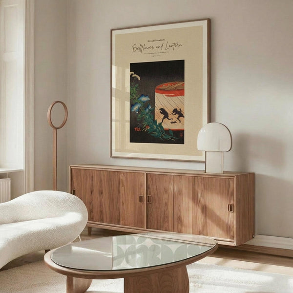 May include: Framed Japanese woodblock print featuring bellflowers and a red lantern displayed above a walnut credenza in a minimalist living room. The artwork shows dark silhouettes against cream matting in a thin wood frame. Modern white sculptural chair and glass oval coffee table complete the serene, neutral-toned space with natural lighting.