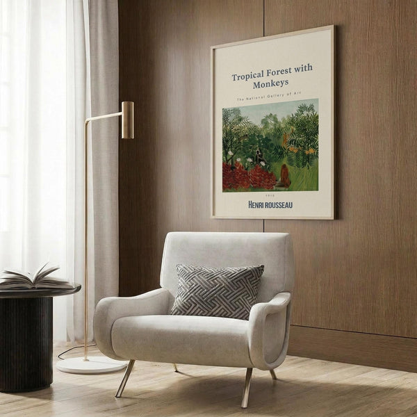 May include: cream-framed art print displaying tropical jungle scene with lush green foliage and small figures, mounted on wood-paneled wall above beige upholstered armchair with geometric patterned pillow, brass floor lamp with white marble base, and dark round side table with open book near window with light curtains