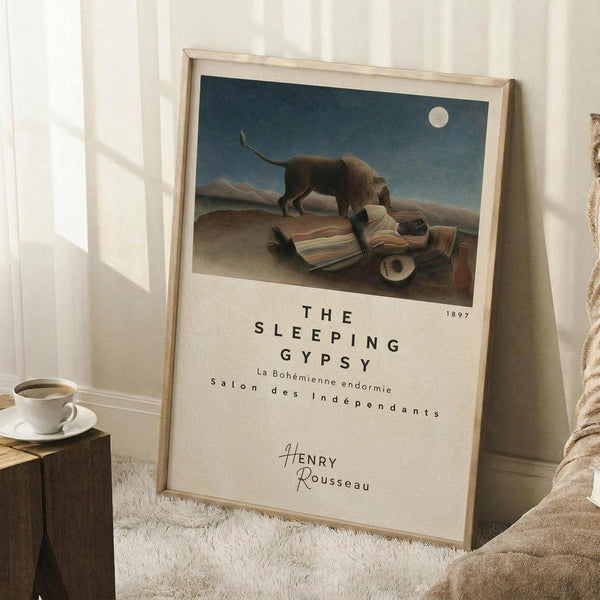 May include: Framed art print leaning against white wall showing Henri Rousseau's famous painting of a sleeping figure in striped robes beside a lion under moonlight in desert landscape. Light wood frame, cream matting with black text below artwork. Cozy room setting with white coffee cup on wooden block, cream throw blanket, and soft natural lighting creating shadows on textured rug.