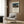May include: Cream-colored wall with wooden paneling displaying a framed art print featuring a sleeping figure under moonlight with an elephant, alongside a beige upholstered armchair with geometric patterned pillow, brass floor lamp with white shade, and dark round side table with open book