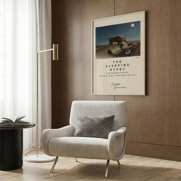 May include: Cream-colored wall with wooden paneling displaying a framed art print featuring a sleeping figure under moonlight with an elephant, alongside a beige upholstered armchair with geometric patterned pillow, brass floor lamp with white shade, and dark round side table with open book