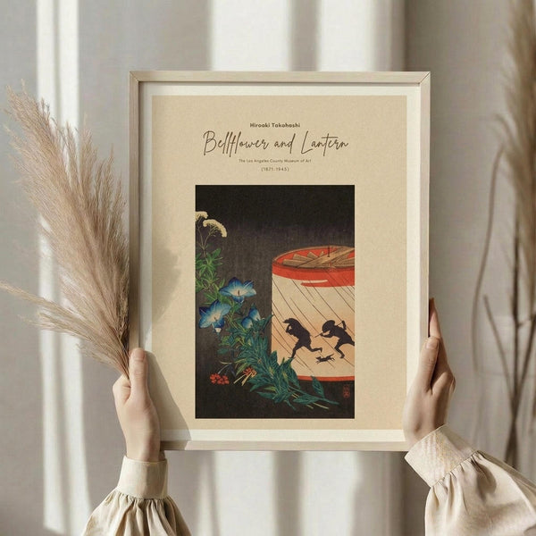May include: Person in beige ruffled sleeves holding framed vintage Japanese art print featuring blue morning glories, ornamental grasses, and silhouettes of two figures beneath a red paper lantern against dark background. Dried pampas grass held in left hand, neutral curtained background, beige mat and light wood frame.