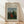 May include: Framed art print leaning against white wall on light wood floor. Print shows woman in red dress sitting in lush green landscape with church spire in background. Cream matting surrounds colorful naive-style painting in thin wooden frame. Soft natural lighting creates diagonal shadows across white textured wall.