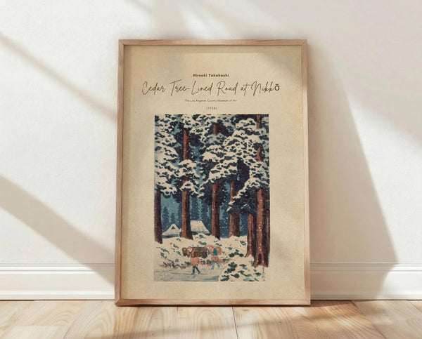 May include: Framed vintage Japanese woodblock print poster featuring snow-covered cedar trees with small figures walking along a snowy mountain path. The artwork shows tall brown tree trunks with white snow-laden branches against a blue winter sky. The cream-colored poster has black text at the top identifying the artist and artwork details. The natural wood frame sits on light wooden flooring against a white wall with soft natural lighting casting gentle shadows.