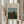May include: Person in cream ruffle-sleeve top holding framed art print showing woman in red dress sitting in lush green landscape with church spires in background. Natural wood frame, cream matting. Dried wheat stalks visible on left. Soft natural lighting against white curtained window.